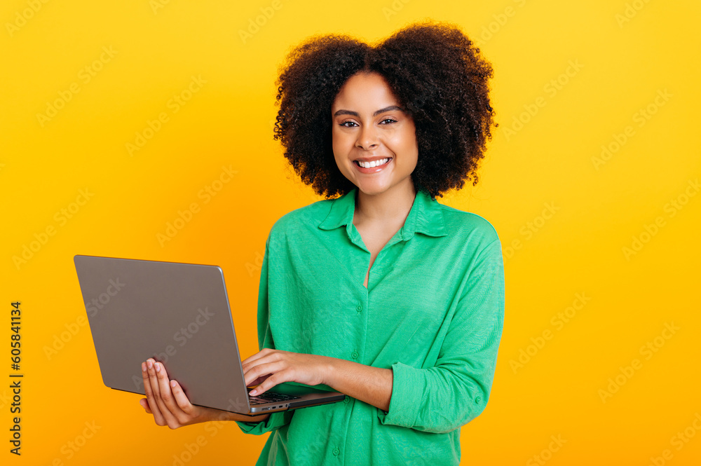 Positive beautiful brazilian or african american curly haired woman, dressed in a green shirt, holding an open laptop in hand, looking at camera, smiling, standing over isolated orange background Positive beautiful brazilian or african american curly haired woman, dressed in a green shirt, holding an open laptop in hand, looking at camera, smiling, standing over isolated orange background