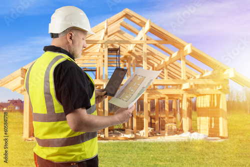 Architect with blueprints. Man is building house. Engineer with tablet. Construction house from boards. Frame building. Frame cottage near architect. Experienced man architect builds country house