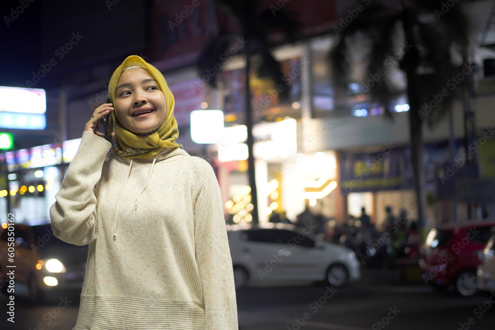 Beautiful Asian Hijab Moslem Young Woman Talking on Smartphone Walking Through Night City Street Full of Neon Light. Portrait of Gorgeous Smiling Female Answering Mobile Phone in Stylish Urban Area. Beautiful Asian Hijab Moslem Young Woman Talking on Smartphone Walking Through Night City Street Full of Neon Light. Portrait of Gorgeous Smiling Female Answering Mobile Phone in Stylish Urban Area.