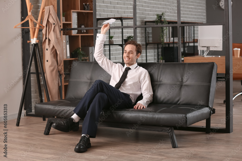 Young businessman with paper plane sitting on sofa in office Young businessman with paper plane sitting on sofa in office