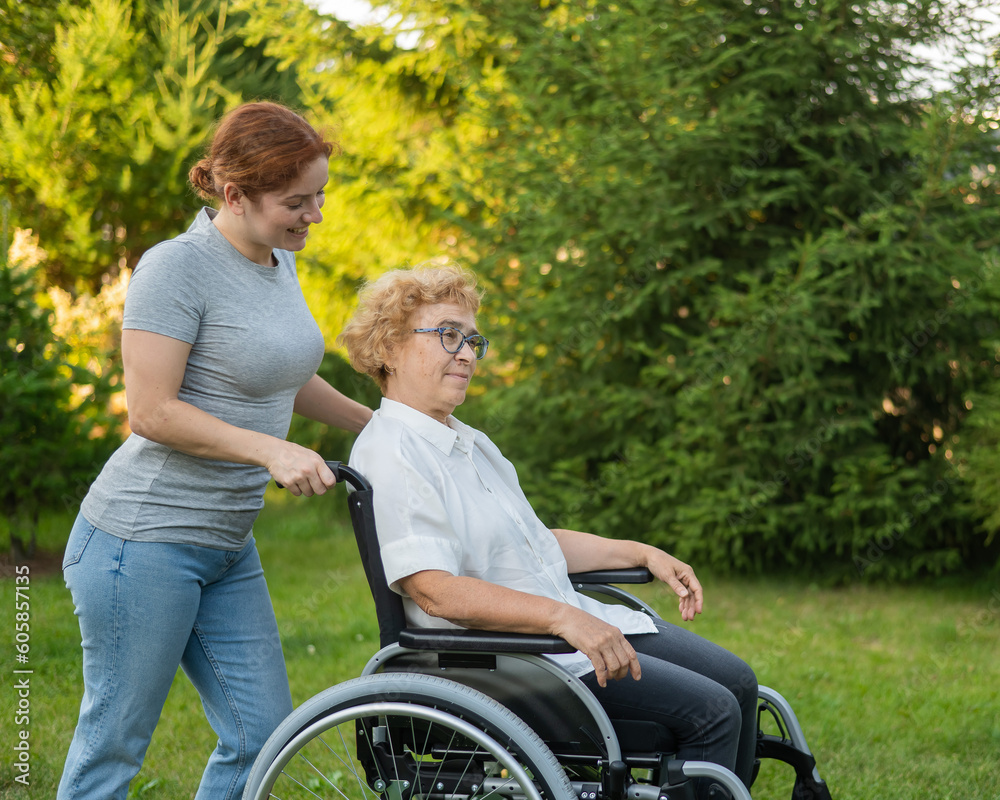 A caucasian woman is carrying an elderly mother sitting in a wheelchair through the park. Walk outdoors. 