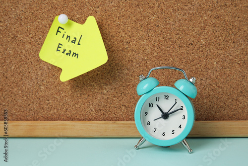 Sticky note with text FINAL EXAM hanging at cork board and alarm clock on blue table