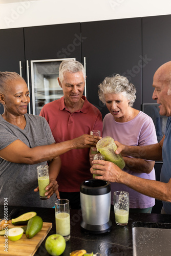 Happy diverse senior friends talking and pouring healthy smoothies together in kitchen