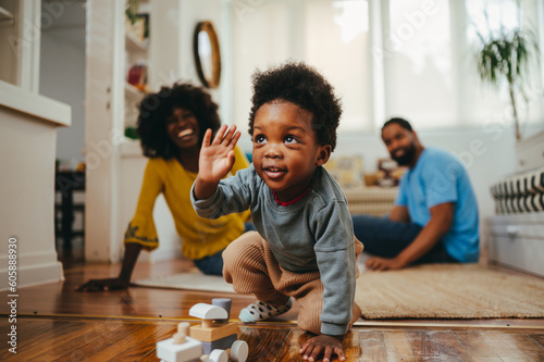 A happy toddler is playing with a wooden educational Montessori train toy on the floor at home while his parents laughing at him from a blurry background.