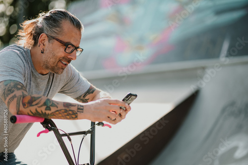 A tattooed middle-aged urban man is sitting on his bmx bike in a skate park and watching at videos on social media.