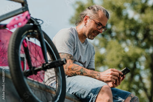 A happy mature tattooed urban man is sitting in a skate park next to his bmx bike and smiling at videos on his mobile phone.