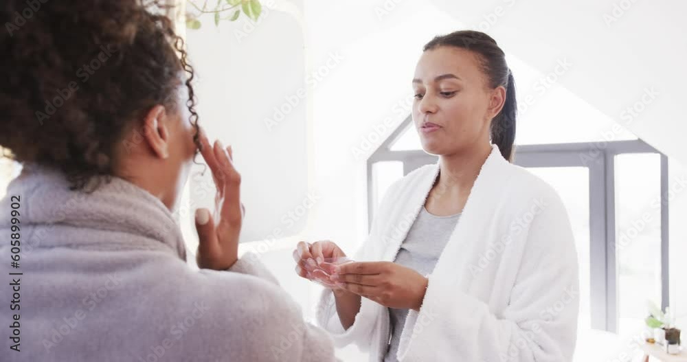 Happy biracial lesbian couple in bathrobes applying under eye masks in sunny bathroom, slow motion Happy biracial lesbian couple in bathrobes applying under eye masks in sunny bathroom, slow motion