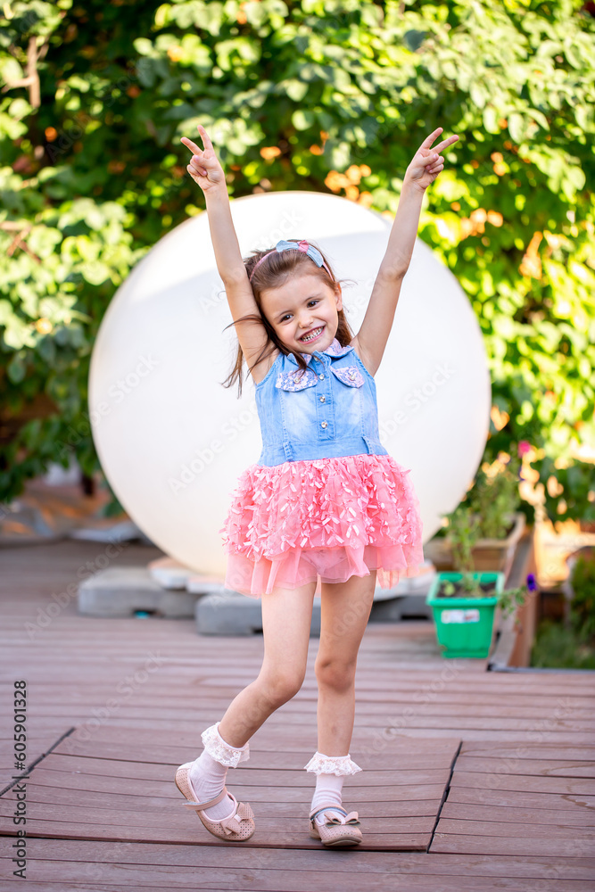 Naklejka premium A cheerful little girl plays in the park on the lawn in a beautiful dress rejoices in the summer. Beautiful white girl smiling while walking in the courtyard of the house.
