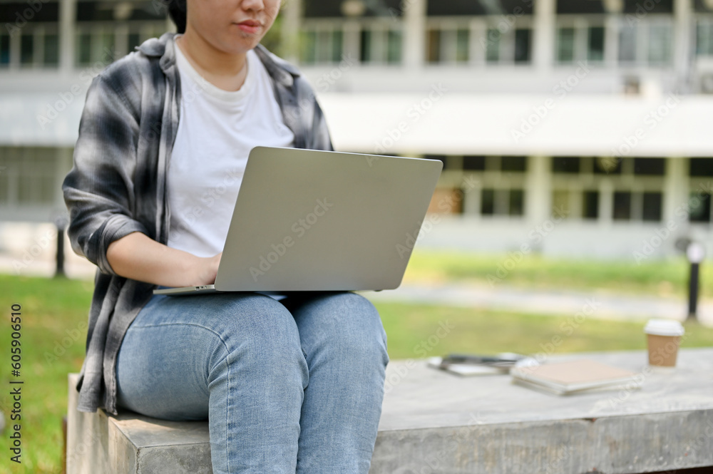 A female college student using her portable laptop on a bench in a campus park.