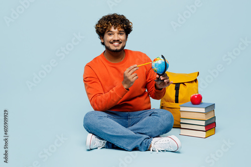 Full body fun young teen Indian boy student wear casual clothes sit near backpack bag pile of books point on globe map isolated on plain pastel blue background. High school university college concept. © ViDi Studio