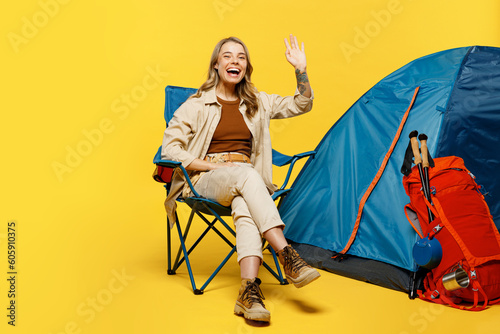 Full body smiling cheerful young woman sit near bag stuff tent waving hand isolated on plain yellow background. Tourist leads active lifestyle walk on spare time. Hiking trek rest travel trip concept,