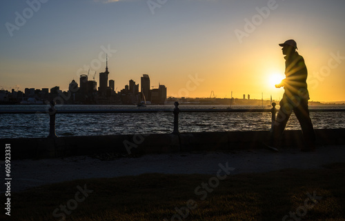 Man walking at Devonport waterfront at sunset. Skytower and Auckland city high-rises in the background.