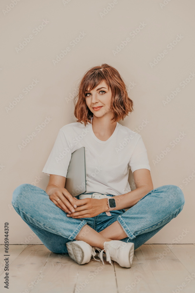 A brunette sits on the floor with a laptop on a beige wall background. She is wearing a white T-shirt, jeans and white sneakers. A brunette sits on the floor with a laptop on a beige wall background. She is wearing a white T-shirt, jeans and white sneakers.