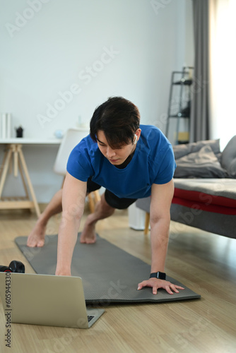 Muscular athletic man in sportswear working out at home and watching fitness lessons online on laptop © Prathankarnpap