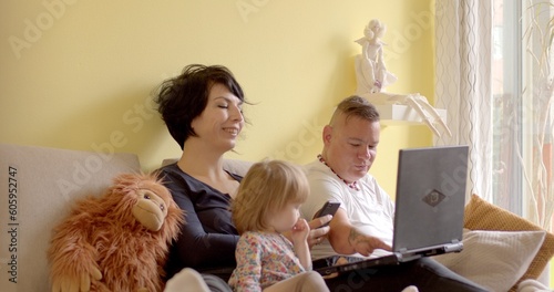 Inspired family is sitting on the sofa in the room. They are using a laptop, the child girl looks into the monitor. The girl presses the buttons. Yellow wall.