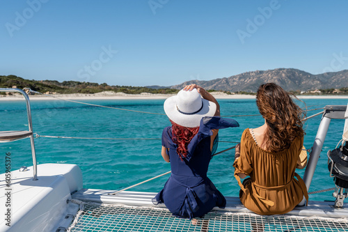 Back view of unrecognizable females in dresses and sunhats sitting on net of sailing yacht on sunny day during summer holiday © Pintau Studio