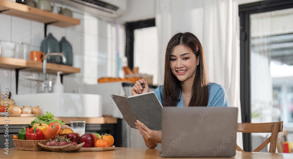 young woman is using laptop computer for remote work or studying online, she takes notes watching webinars or classes at kitchen table at home young woman is using laptop computer for remote work or studying online, she takes notes watching webinars or classes at kitchen table at home