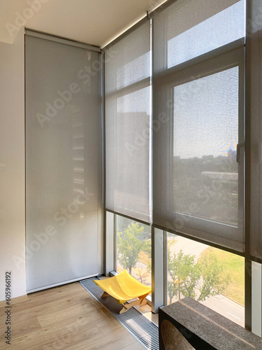 Roller blinds on the windows in the interior. Automatic solar shades on full height windows. Motorized sunscreen blinds with screen fabric. Ginger cat lies on a couch near the window with curtains.