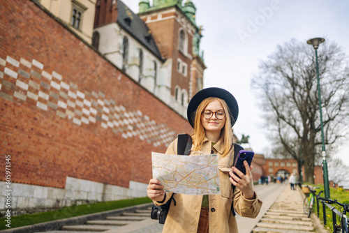 Stylish clothes blond girl tourist travels looking for directions on map and phone while traveling through the old town of Krakow in spring, Poland. Spring view of Wawel Royal Castle  © Shi 