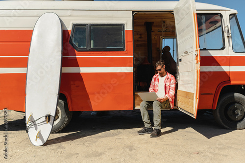 Full body of adult male freelancer in casual clothes and sunglasses working remotely on laptop sitting on van step during road trip photo