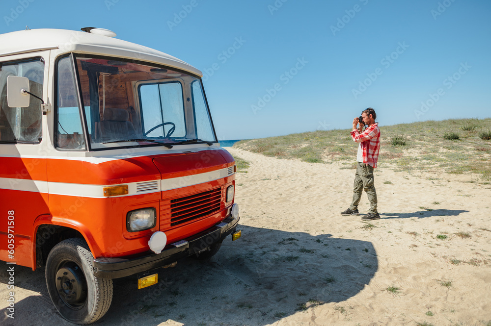 Side view of concentrated male traveler in casual clothes taking photo of sea and sandy beach standing near retro van against cloudless blue sky