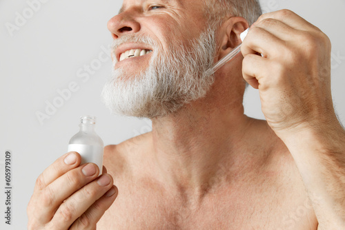 Fototapeta Naklejka Na Ścianę i Meble -  Cropped image of smiling, handsome mature man posing shirtless, applying face serum. Model against grey background. Concept of male beauty, face and skin care, daily procedures, age