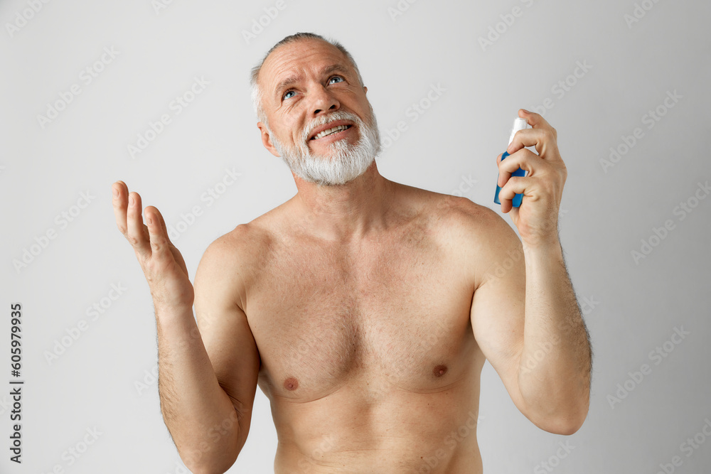 Portrait of handsome, smiling, mature man with gray hair and beard posing shirtless, applying with face toner against grey background. Concept of male beauty, face and skin care, daily procedures, age