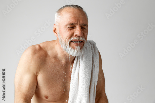 Portrait of mature, grey-haired, shirtless man doing morning routine, washing face, taking care after skin against grey background. Concept of male beauty, face and skin care, daily procedures, age