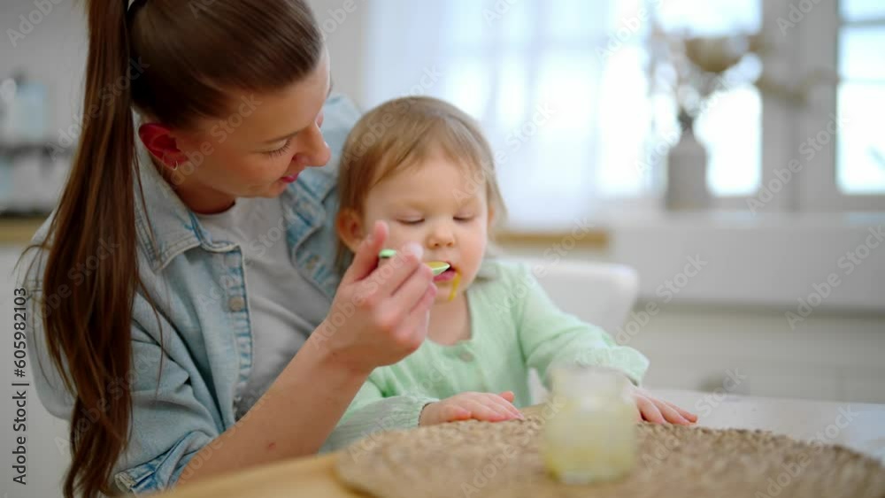 Mother feeds her baby fruit or vegetable puree using plastic spoon. Toddler eats with pleasure. Mom holds child on knees at table on kitchen. Raising child, kid's nutrition and motherhood concept.