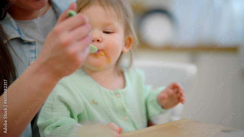 Mom feeds her baby playing with food. Toddler eats fruit or vegetable puree. Mother holds child on knees at table on kitchen. Raising child, kid's nutrition, motherhood, love care in family concept. Mom feeds her baby playing with food. Toddler eats fruit or vegetable puree. Mother holds child on knees at table on kitchen. Raising child, kid's nutrition, motherhood, love care in family concept.
