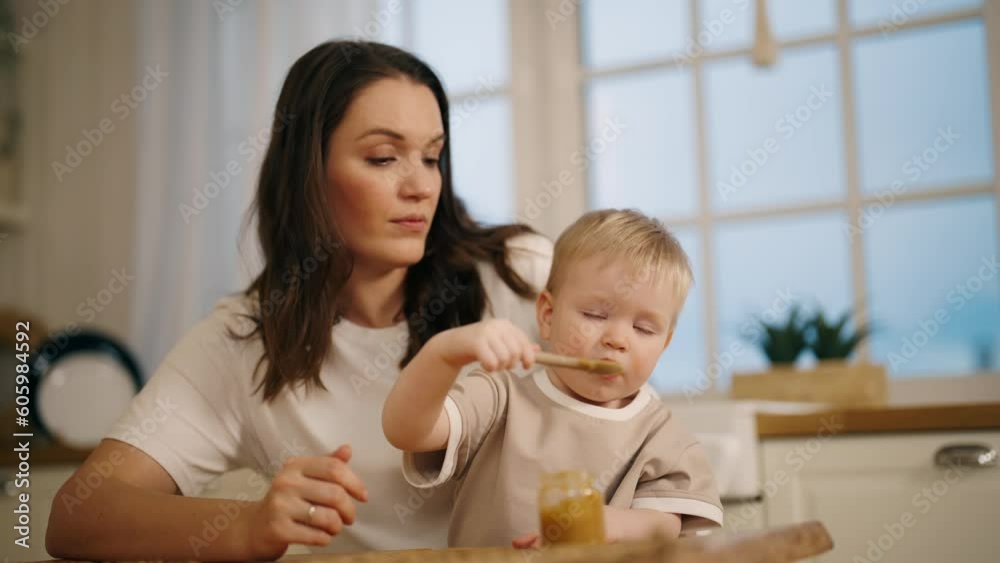 Happy smiling positive mom looks on baby girl eats herself fruit or vegetable puree from jar with spoon sits on kitchen at home. Babyhood, childhood, maternity leave, rising child motherhood concept. Happy smiling positive mom looks on baby girl eats herself fruit or vegetable puree from jar with spoon sits on kitchen at home. Babyhood, childhood, maternity leave, rising child motherhood concept.