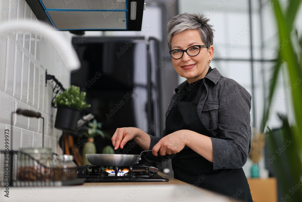 Φωτογραφία An older woman in glasses and an apron is frying food in a frying pan in the kitchen