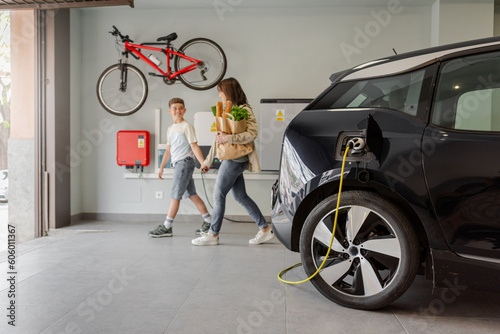 Fotografia, Obraz Electric vehicle charging station in private home with happy mother and son walking alongside, blurred family leaving the house, with a bicycle hanging on the wall