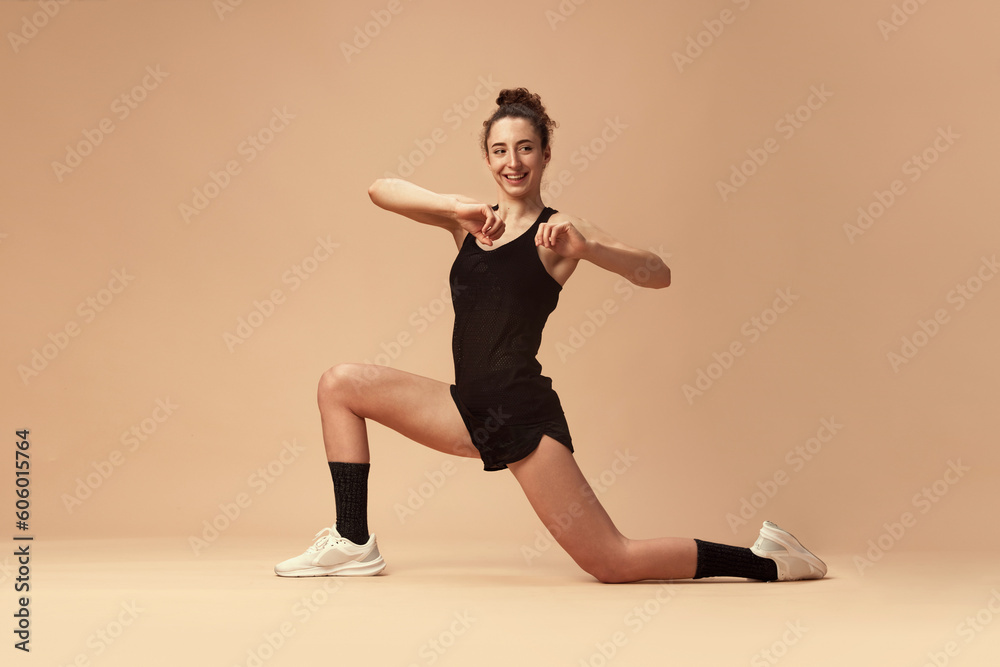 Flexible, sportive, smiling young girl in sportswear training, stretching against light brown studio background. Workout. Concept of sportive lifestyle, beauty, body care, fitness, health