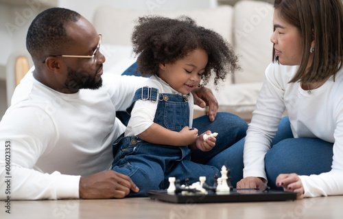 Little kid girl play chess board at home with multiethnic family. Adorable afro hair multiracial daughter sitting on floor enjoy playing piece of chess with dad and Asian mother. Leisure childhood.
