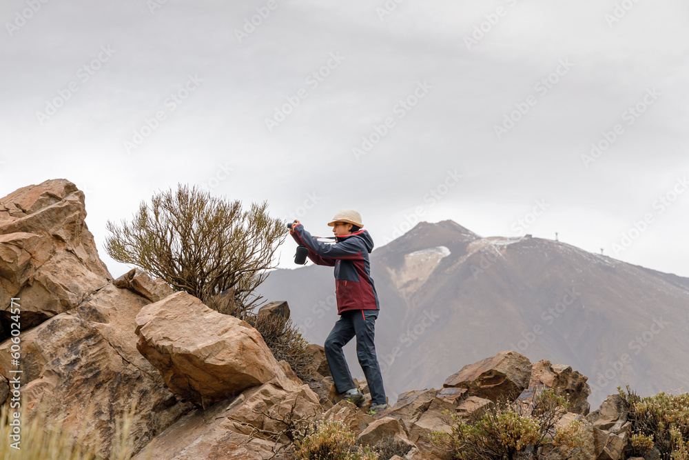 Naklejka premium Teenage girl with a hat taking a picture of a bush, with the Teide volcano in the background, Tenerife