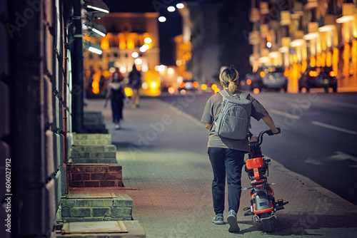 Woman with backpack walk alone on night city street with electric scooter. Girl rolls scooter with dead battery next to her. Night ride on electric scooter. © Tricky Shark