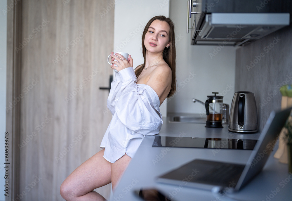 Beautiful Young Girl is Standing in the Kitchen and Drinking Coffee. Morning Routine. Wearing White shirts. Blurry Laptop and Mobile Phone on the Desk. Beautiful Young Girl is Standing in the Kitchen and Drinking Coffee. Morning Routine. Wearing White shirts. Blurry Laptop and Mobile Phone on the Desk.