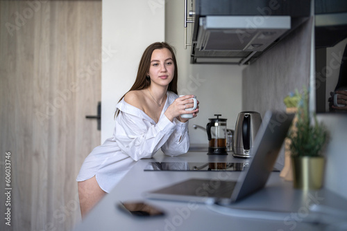 Beautiful Young Girl is Standing in the Kitchen and Drinking Coffee. Morning Routine. Wearing White shirts. Blurry Laptop and Mobile Phone on the Desk.