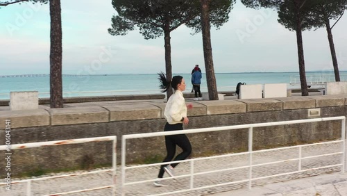 Aerial video of Caucasian woman running waterfront public park. Active runner alone in sportswear exercising outdoor. Healthy lifestyle concept. Portugal, Lisbon, Park of Nations river Tejo Tagus