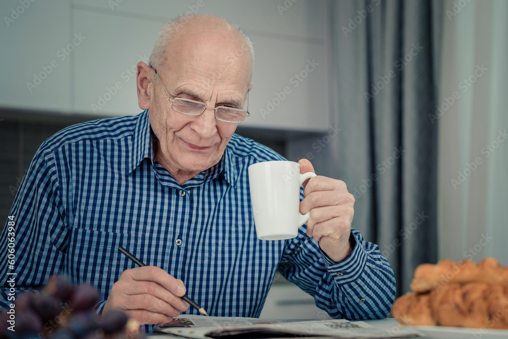 Happy senior man with pen in hand wearing eyeglasses solving crossword and drinking coffee sitting at kitchen table indoor. - obrazy, fototapety, plakaty 
