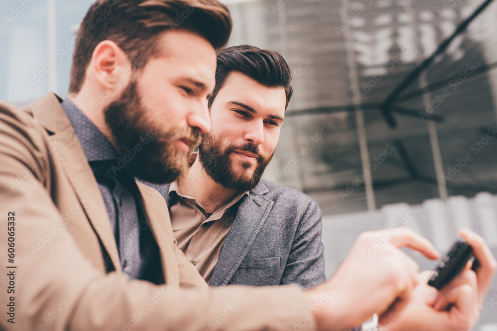 Two casual businessmen outdoors chatting using smart phone Two casual businessmen outdoors chatting using smart phone