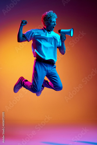 Portrait of mature an in casual clothes jumping and shouting in megaphone against orange studio background in blue neon light. News. Concept of human emotions, lifestyle, youth, facial expression photo