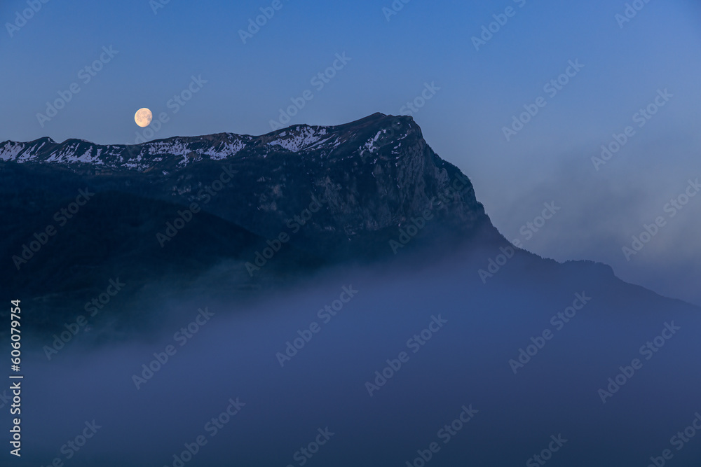 A scenics view of majestic snowy mountain summit (Pic de Morgon) at dawn with a full moon, Hautes-alpes, France under a majestic blue sky