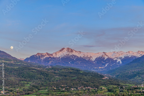 A scenics view of majestic snowy mountain summit (Les Orres) at dawn with a full moon, Hautes-alpes, France under a majestic blue sky © Dolwolfian