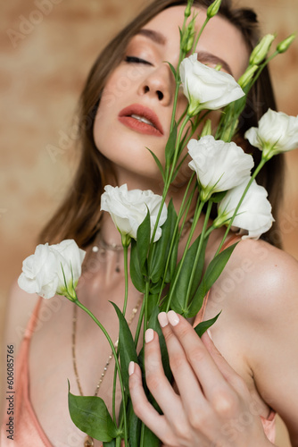 portrait of sensitive and young woman in pink silk slip dress holding eustoma flowers while standing with closed eyes on mottled beige background  sensuality  elegance  sophistication