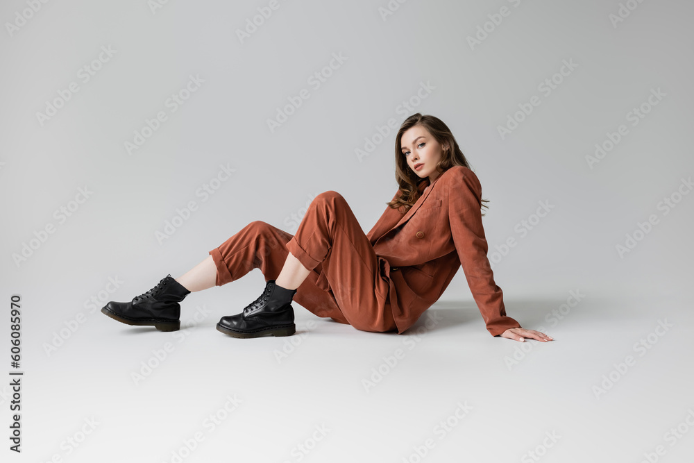 full length of trendy model with brunette and wavy hair sitting in trendy and oversize suit with terracotta blazer, pants and black boots, looking at camera on grey background, stylish young woman - obrazy, fototapety, plakaty 