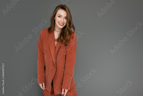 happy young woman with brunette and wavy hair posing in trendy and oversize suit with terracotta blazer and smiling while looking at camera on grey background, stylish pose, trendy chic