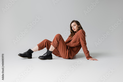 full length of trendy model with brunette and wavy hair sitting in trendy and oversize suit with terracotta blazer, pants and black boots, looking at camera on grey background, stylish young woman