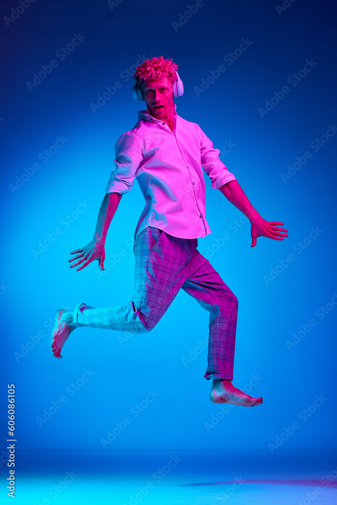 Full-length portrait of mature man listening to music in headphones and jumping against blue studio background in pink neon light. Concept of human emotions, lifestyle, youth, facial expression